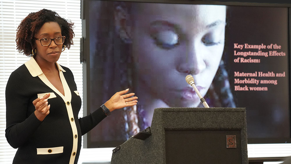 Woman speaking in front of a screen with an image of a Black woman.