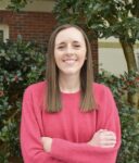 A woman with straight brown hair wearing a pink sweater stands outside with arms crossed, smiling in front of leafy shrubs with red berries.