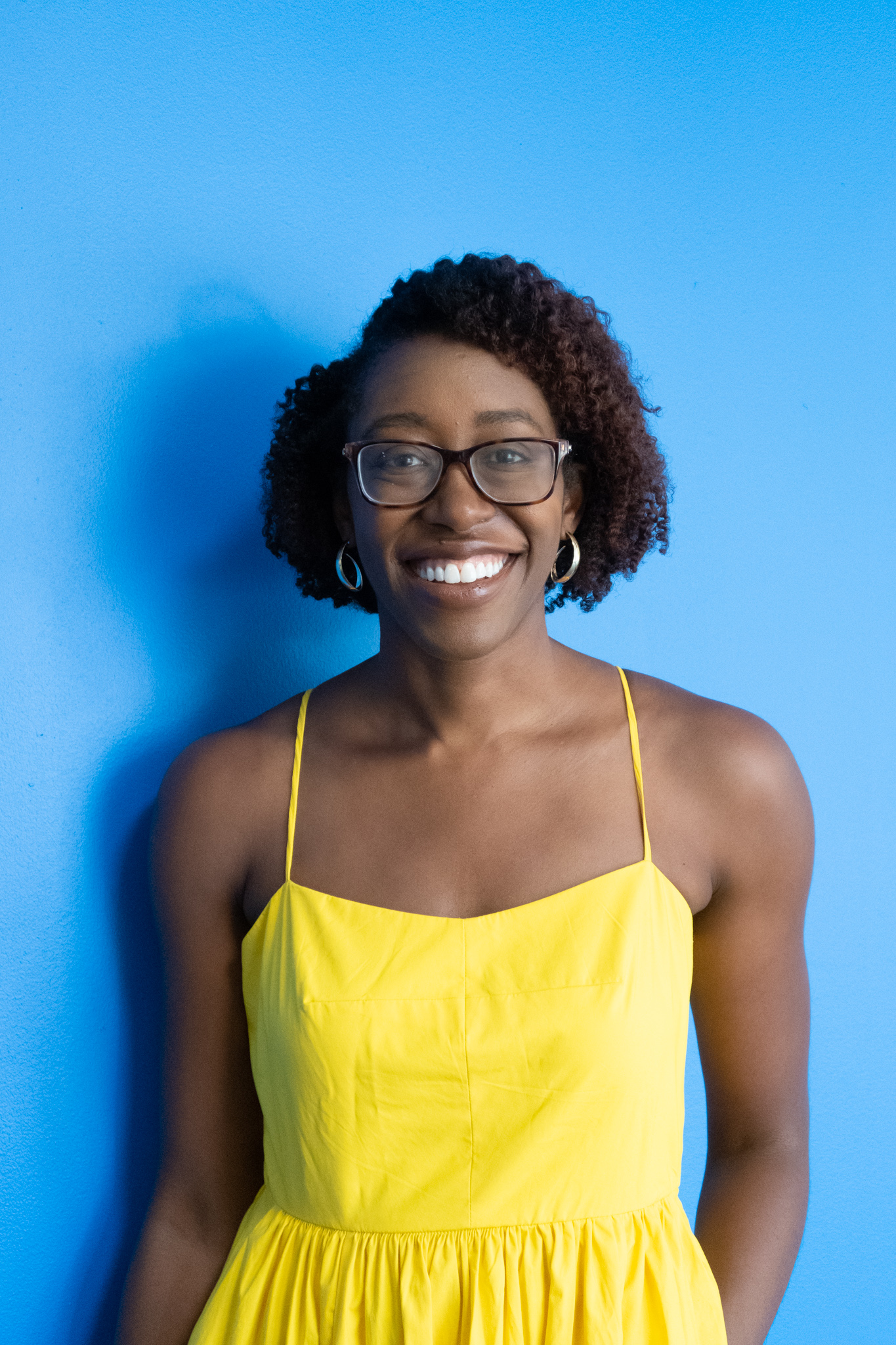 A woman wearing glasses and a yellow dress stands smiling in front of a bright blue wall.
