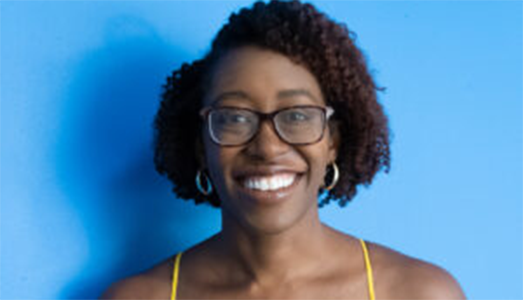 A woman with short curly hair, glasses, and hoop earrings smiles in front of a bright blue background.